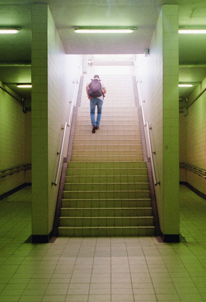 Person with backpack ascending a tiled staircase in Vila Nova de Famalicão subway with soft lighting.