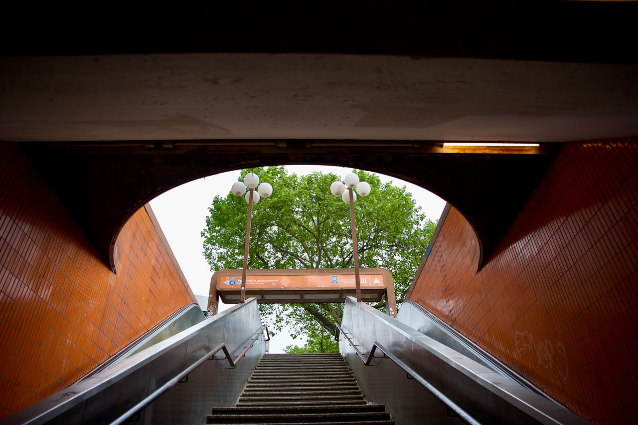 A subway exit featuring an escalator leading outdoors with lamps and greenery in view.